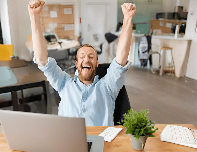 Grinning widely, a white man leans back in his chair, arms raised triumphantly, laptop open in front of him displaying a cheerful message, celebrating a successful achievement.